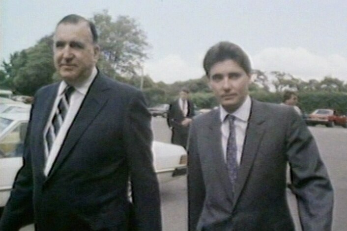 Two men in suits walking along the street, one older and taller, the other younger with brown hair.