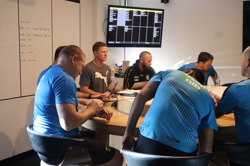 A group of men sit around a table, a game of AFL plays on a TV screen