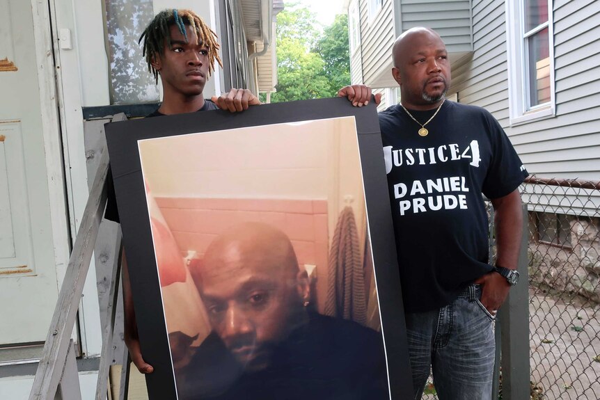 Joe Prude, brother of Daniel Prude, right, and his son Armin, stand with a picture of Daniel Prude at the front of a house.