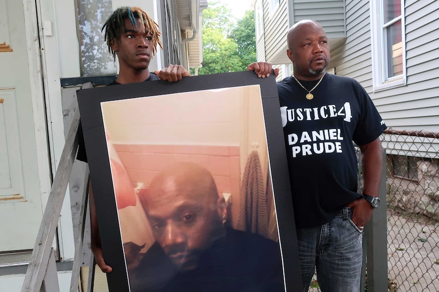 Joe Prude, brother of Daniel Prude, right, and his son Armin, stand with a picture of Daniel Prude at the front of a house.