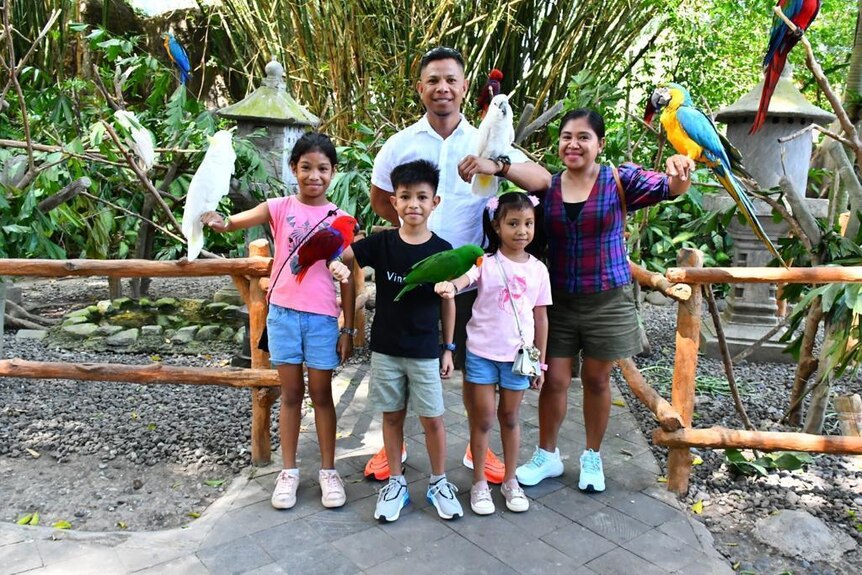 A man stands with his wife, two young daughters and a son, smiling as colourful birds perch on their arms.