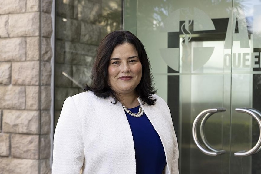 A dark-haired woman in a light blazer stands outside an office building.