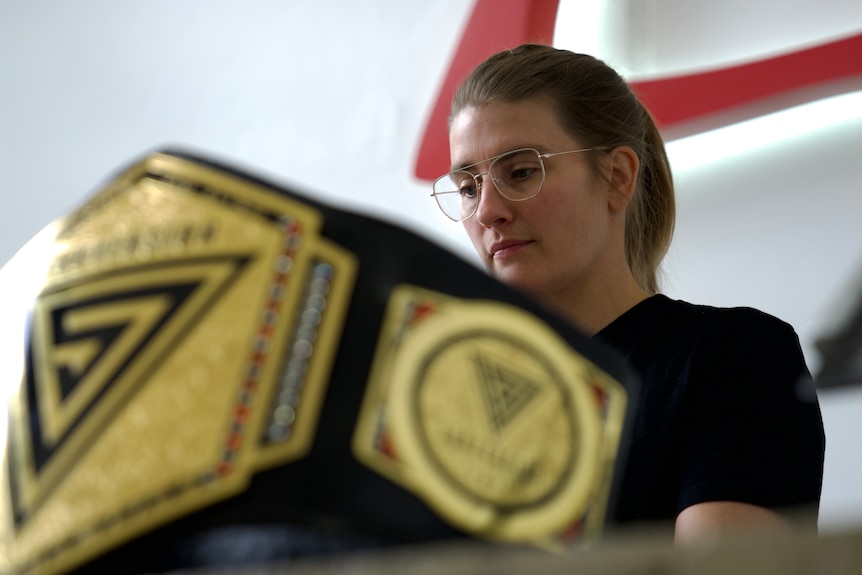 Catherine Wilson sits at a laptop in the background, in the foreground a wrestling belt sits on the table
