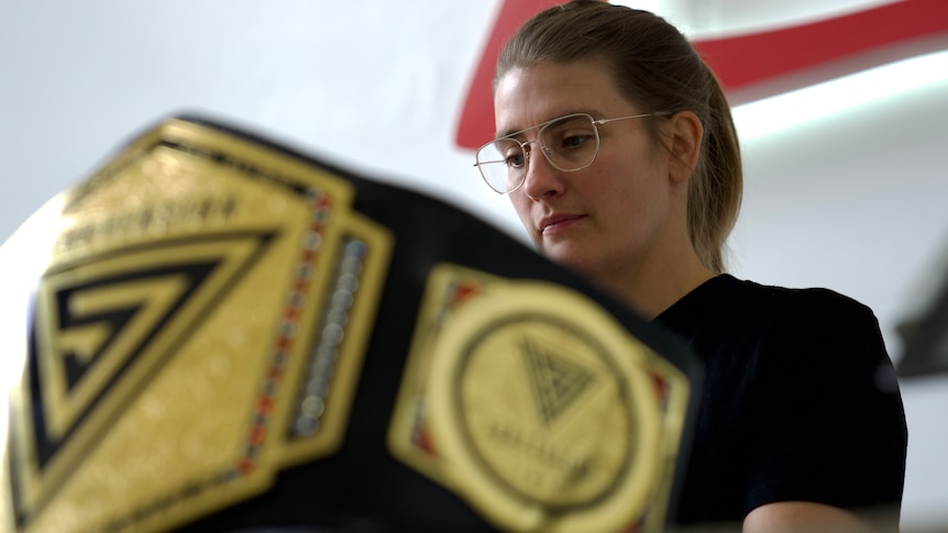 Catherine Wilson sits at a laptop in the background, in the foreground a wrestling belt sits on the table