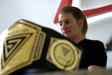 Catherine Wilson sits at a laptop in the background, in the foreground a wrestling belt sits on the table