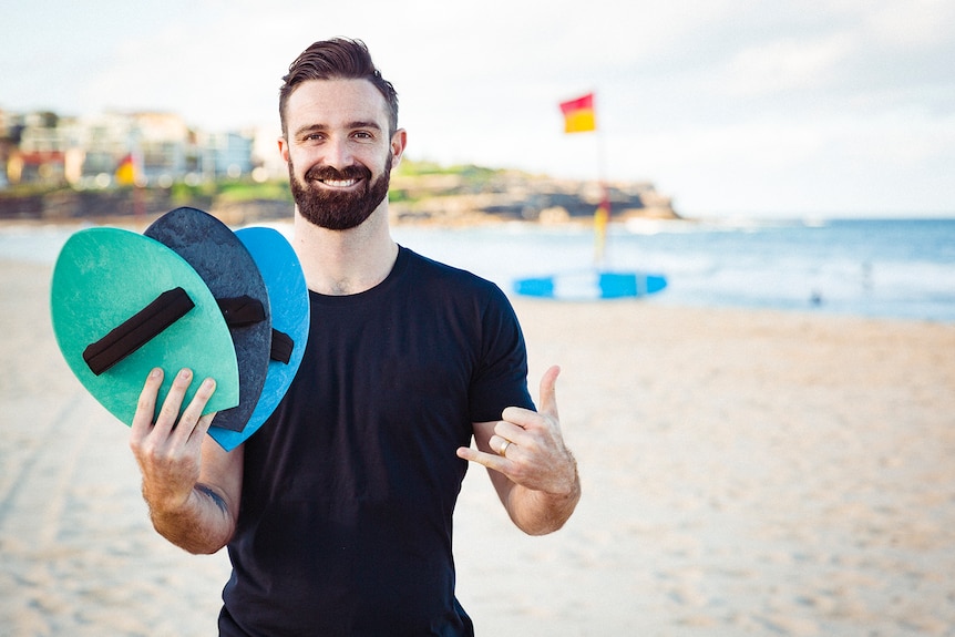 A man holds up three small hand planes with velcro straps, at a beach.