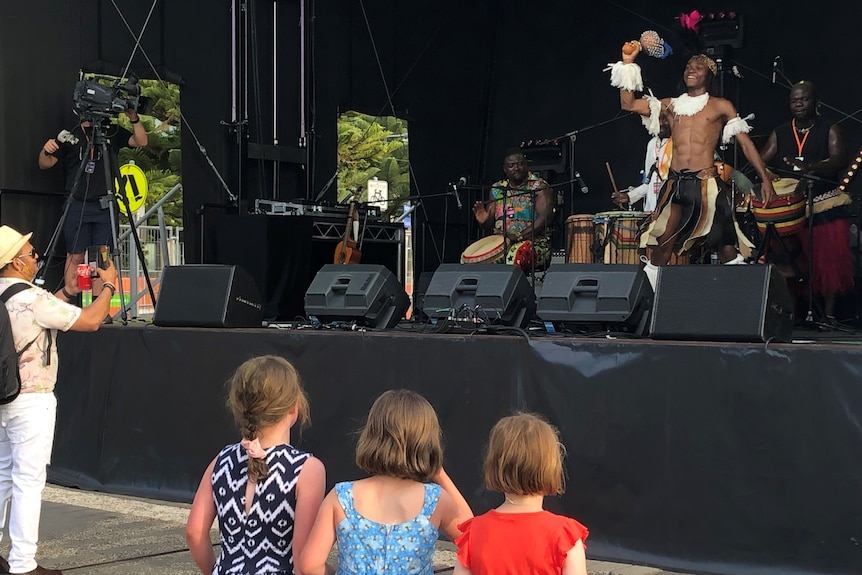 Three small girls stand in front of a stage watching a dancer perform 