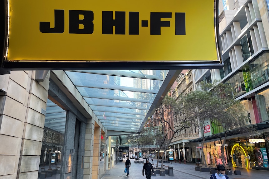 Yellow JB Hi-Fi signage above a fairly empty retail strip, with a shopper walking past wearing a mask.