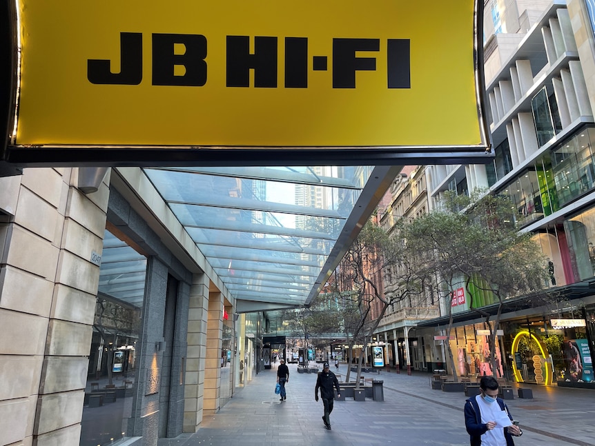 Yellow JB Hi-Fi signage above a fairly empty retail strip, with a shopper walking past wearing a mask.