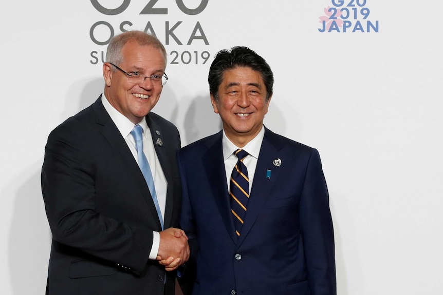 Two men in suits shaking hands in front of a poster reading "G20 2019 Japan"