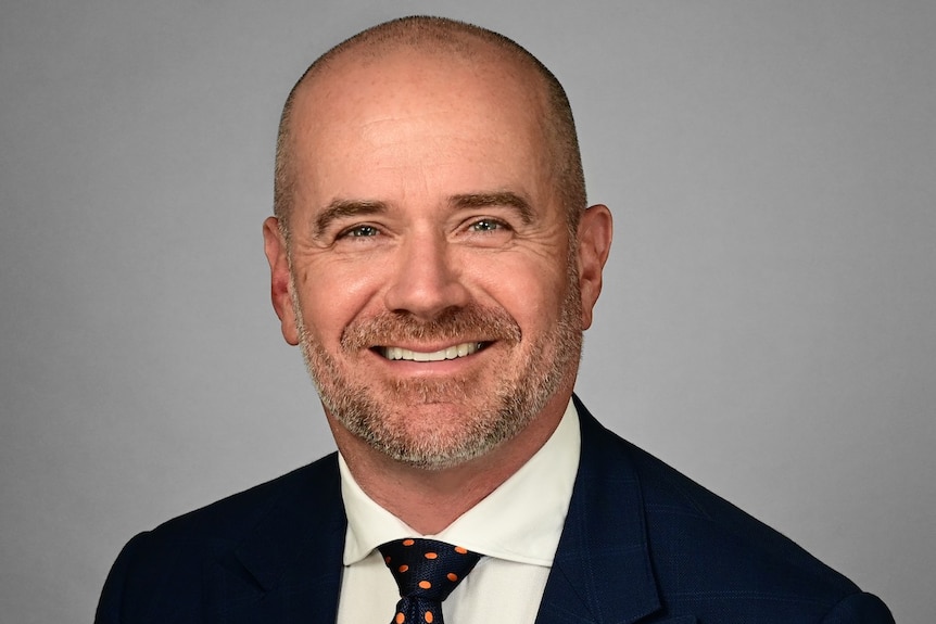 A bald man with grey beard and moustache smiling wearing a black suit, white shirt, red tie