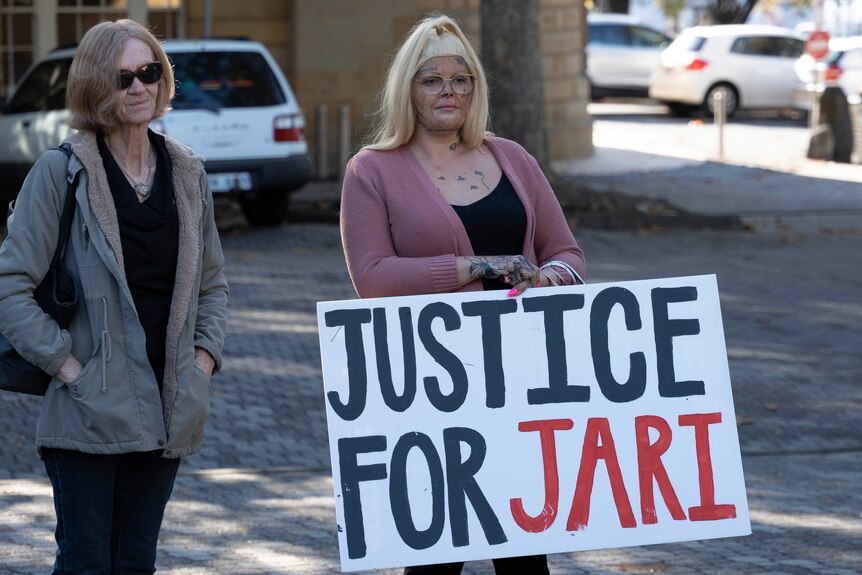 A woman holds a sign saying 'Justice for Jari' outside a court building.
