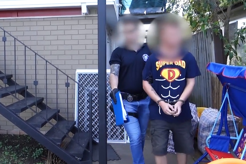 man in 'super dad' t shirt led from a house in shackles, police officer behind him, both faces blurred