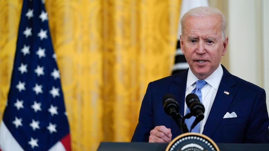 President Joe Biden speaks into micrpohones as he stands new a US flag.