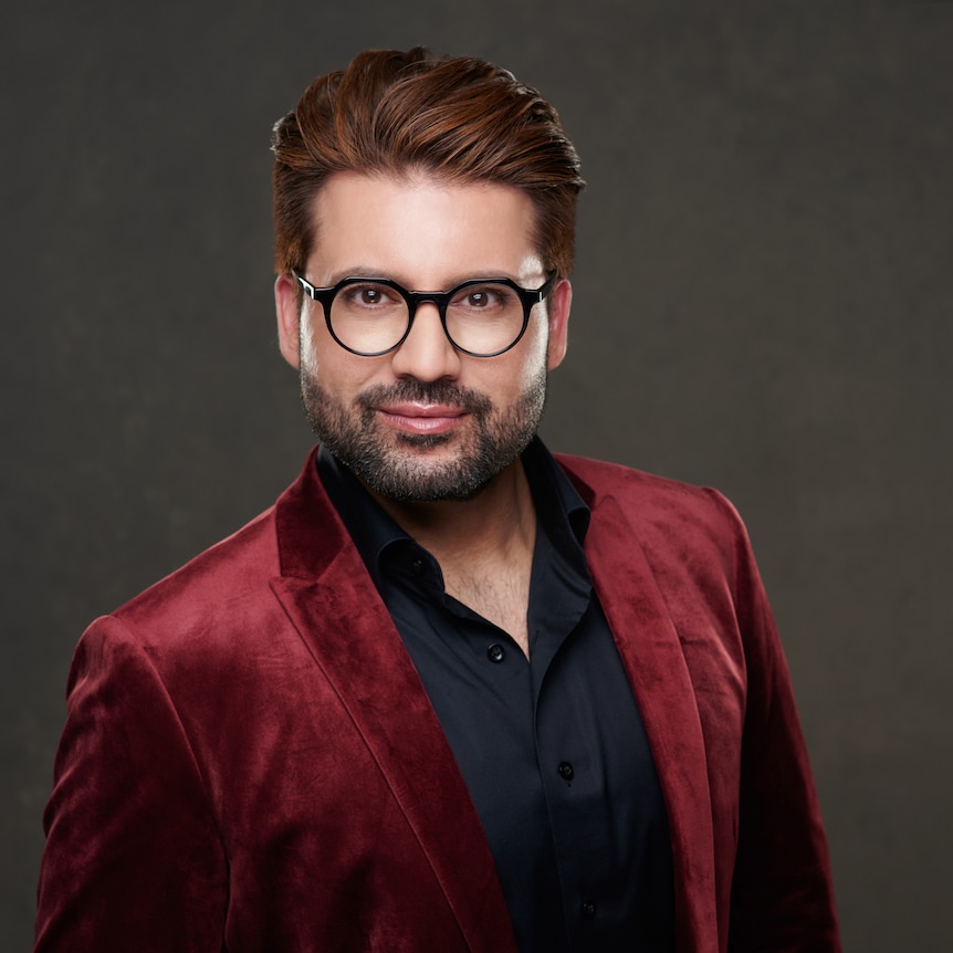 A man wearing a red suit looks at the camera in front of a dark background.