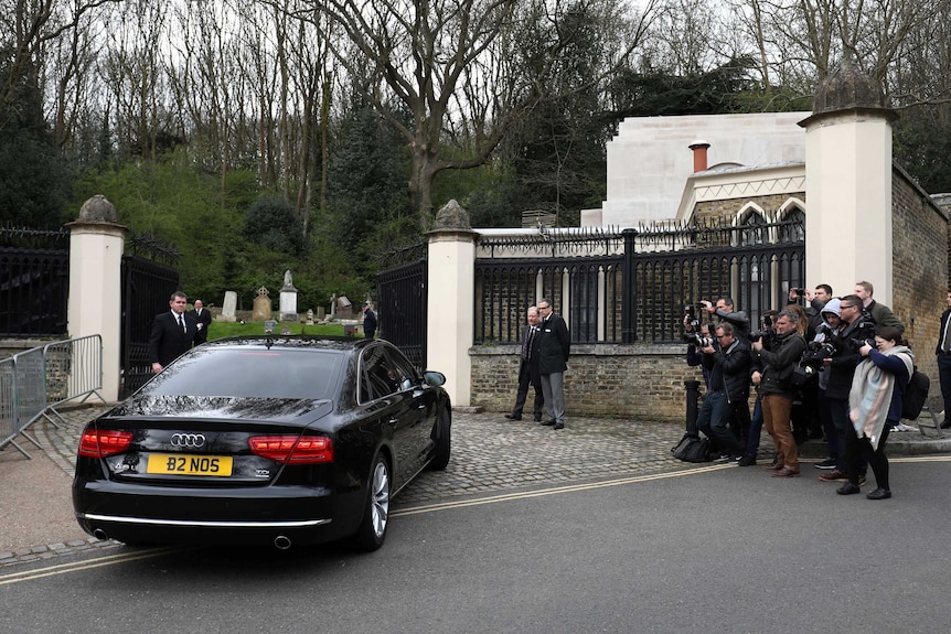 A car drives past media and through gates of Highgate Cemetery in London.