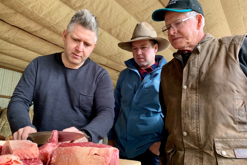 A chef cuts beef on a chopping board, watched by farmers