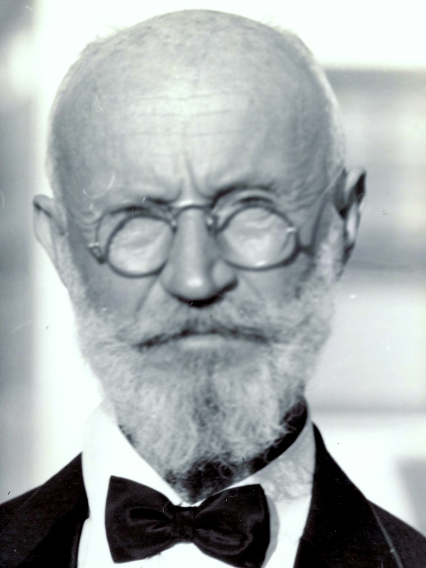 A black and white archival photo of an elderly bald white man with a white beard, spectacles and a bow-tie.