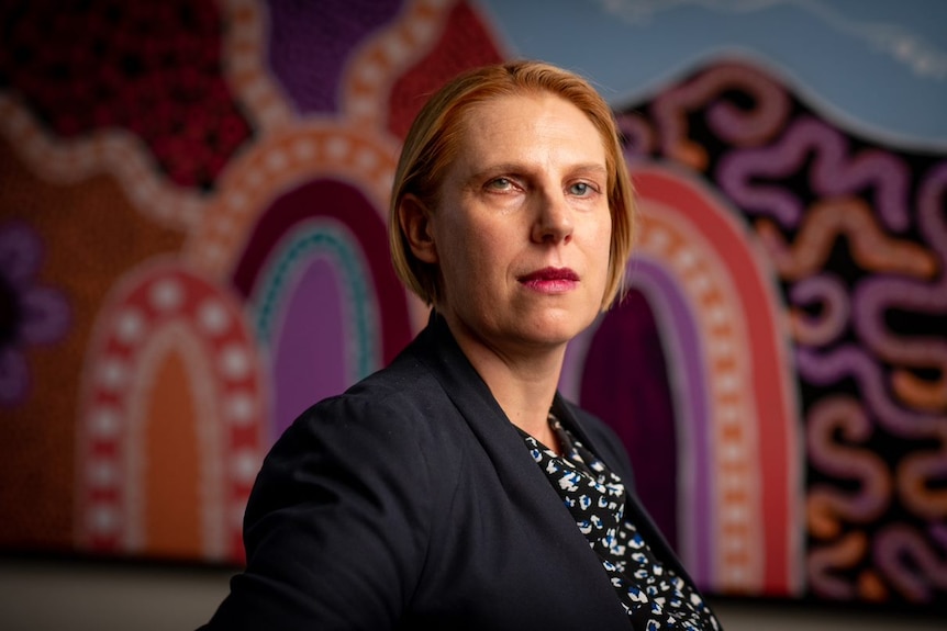 Leah Bromfield looks stern with Aboriginal painting behind her.