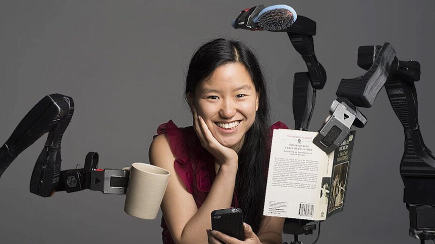 Marita is smiling and sitting between two robot hands holding items.