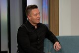 TV psychic John Edward sits on a blue couch in a TV studio. He wears a black button-up shirt and is looking off screen.