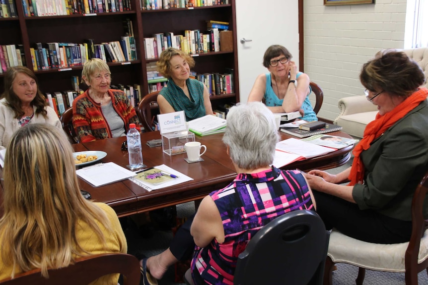 Seniors sitting around a table listening to another senior talking with bookcase full of books in background.