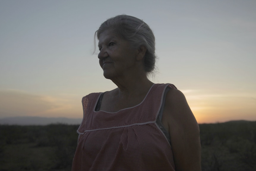 Twilight shot of an older woman with grey hair in bun, wearing cheesecloth smock, with arid landscape in background.