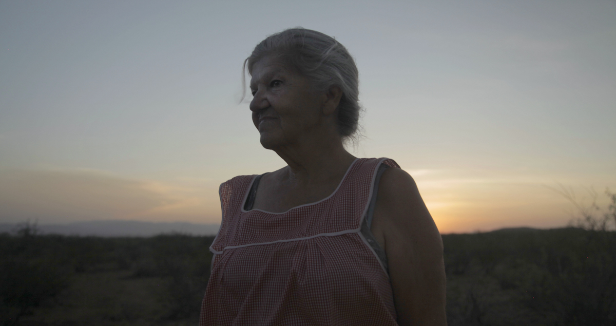 Twilight shot of an older woman with grey hair in bun, wearing cheesecloth smock, with arid landscape in background.