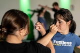 Two young women cross arms, hands held in tight fists, while others appear in hand-to-hand combat in the background.