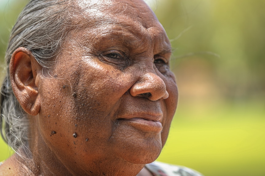 A close up of an elderly Indigenous woman looks away from the camera with a neutral facial expression.