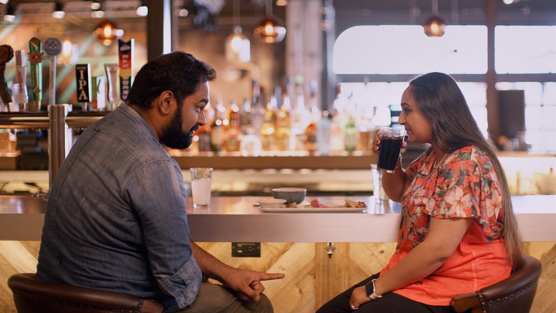A young Indian man with a denim shirt sits at a bar next to a young Indian woman who is sipping coca-cola.