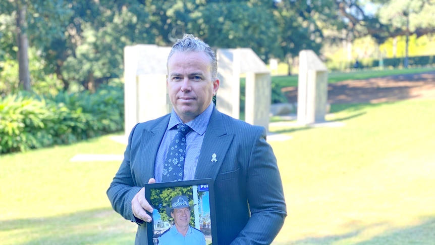 Man standing with a photo of an older man, his father in police uniform, in a garden setting.