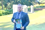 Man standing with a photo of an older man, his father in police uniform, in a garden setting.