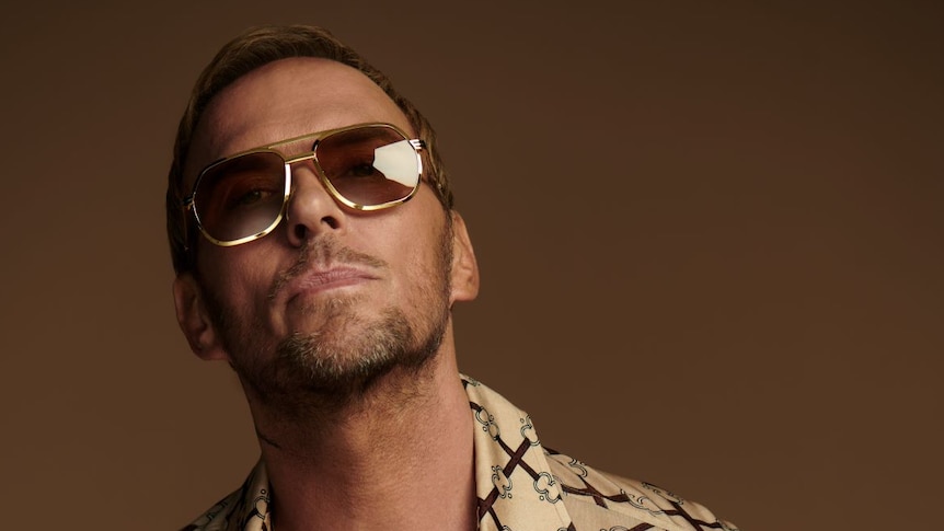 Bros star Matt Goss wearing metal-rimmed sunglasses and a brown shirt unbuttoned to show tattoos on his chest.