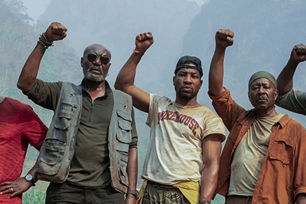 On hazy day in mountainous jungle six men  with serious expressions wear hiking attire and stand with raised right fists.