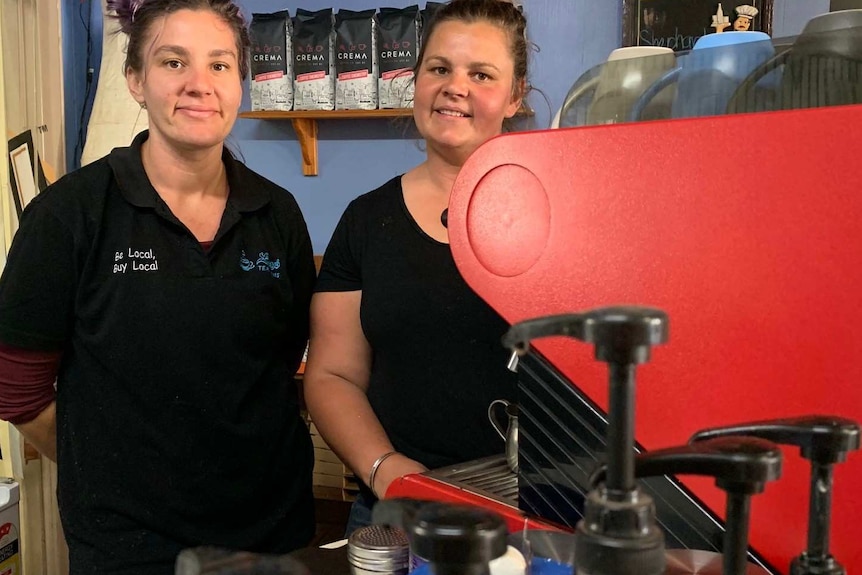 Amy and Jess stand behind the counter of their cafe at the red coffee machine. The walls are a blue/purple.