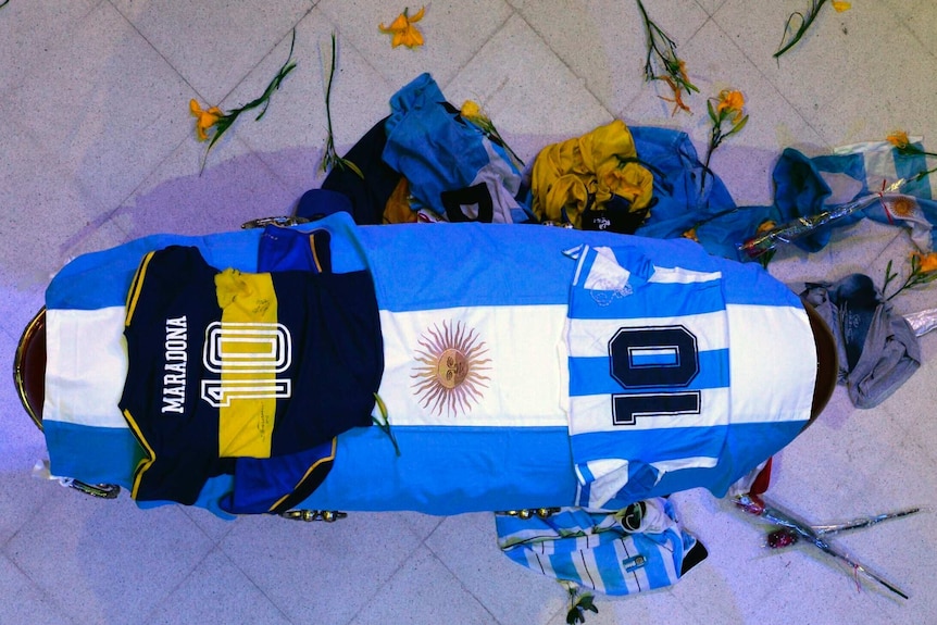 A coffin surrounded by flowers and draped with an Argentina flag and jerseys of Boca Juniors and Argentina.