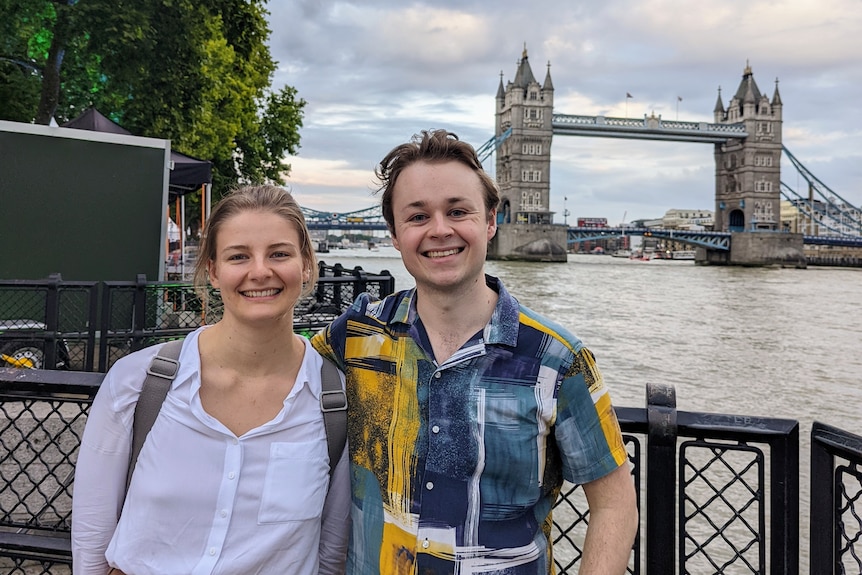 A young woman and man stand in London in front of a prominent bridge and river.