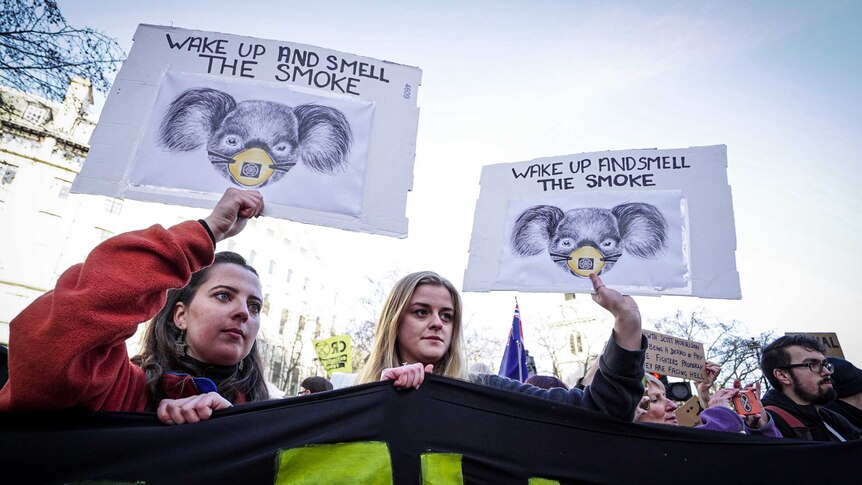 Two women hold up signs showing koalas wearing face masks reading "wake up and smell the smoke"
