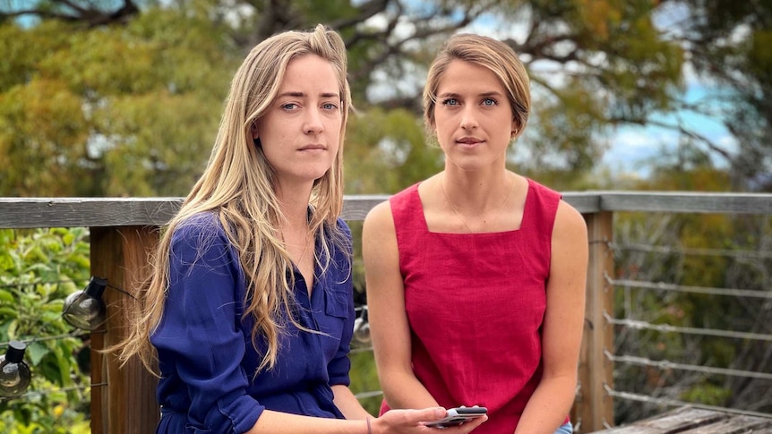 Two women sit next to each on a bench looking at the camera.