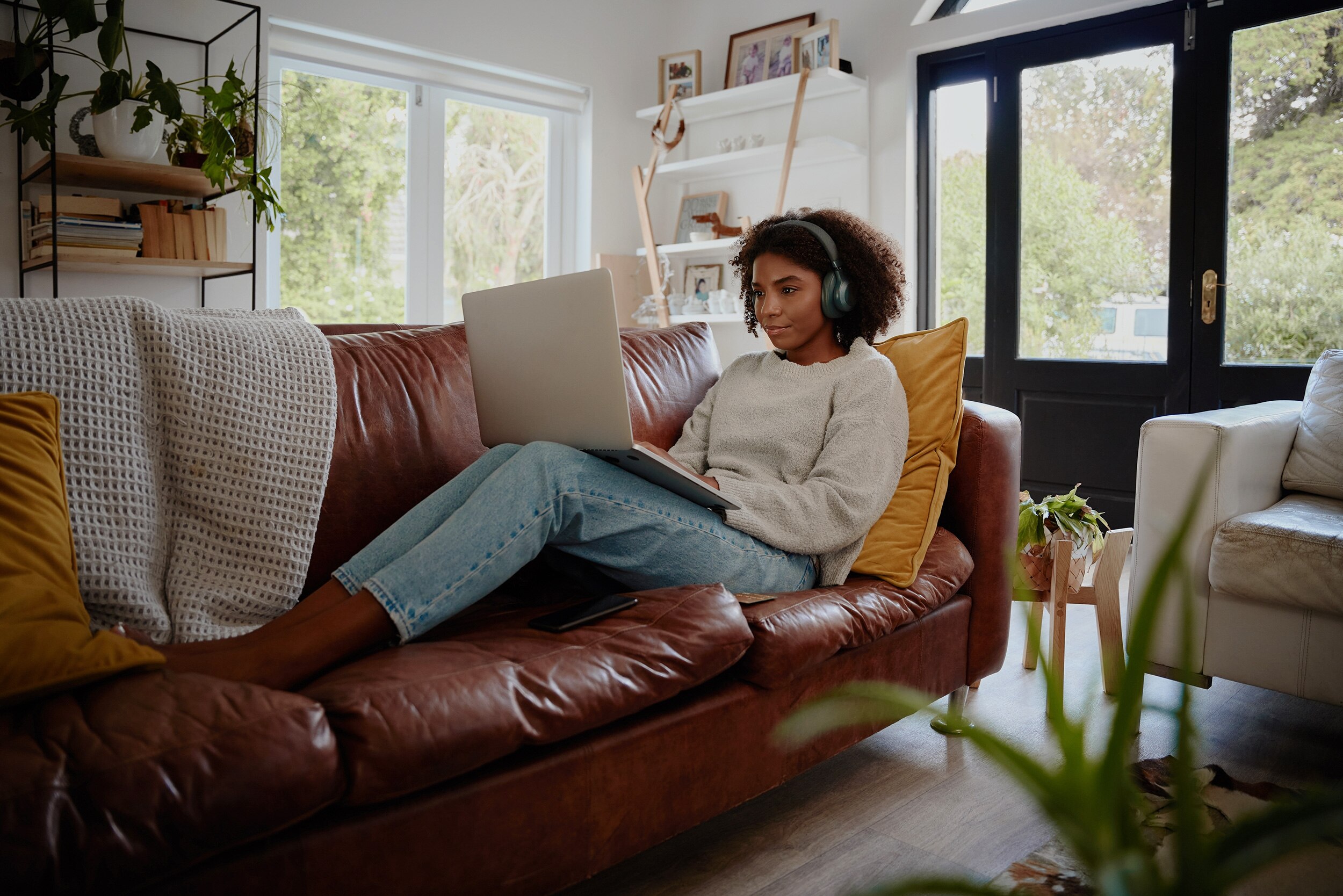 A woman with curly hair sits on a couch with a laptop on her legs and headphones on.