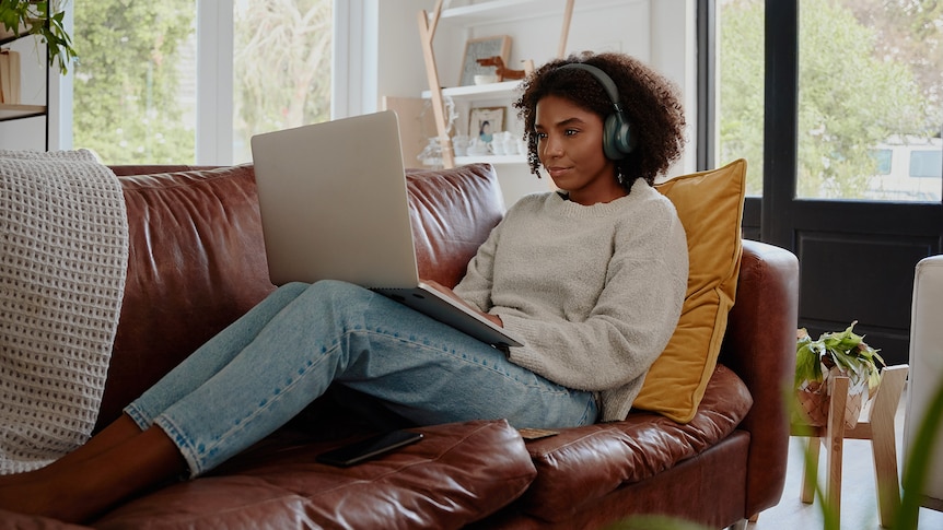 A woman with curly hair sits on a couch with a laptop on her legs and headphones on.