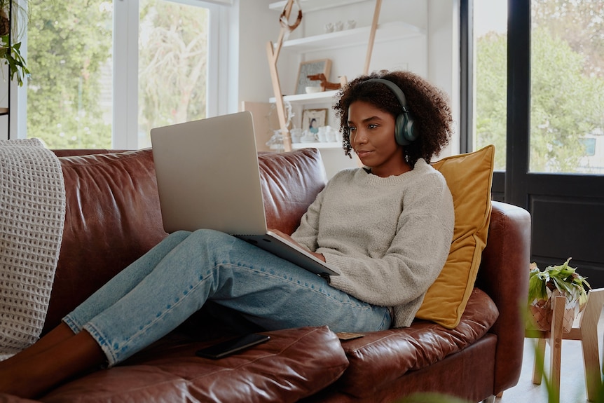 A woman with curly hair sits on a couch with a laptop on her legs and headphones on.