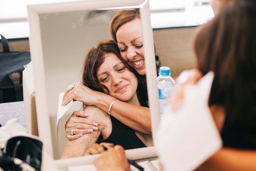 Two women embrace in front of a mirror