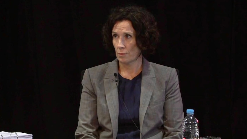woman with short brown hair in grey blazer giving inquiry evidence in front of a stand and microphone