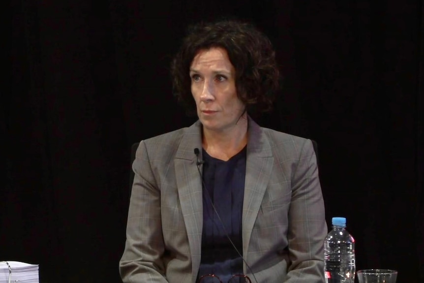 woman with short brown hair in grey blazer giving inquiry evidence in front of a stand and microphone