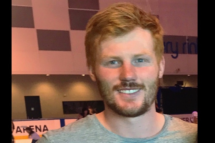 A man with red hair and a beard in a grey top smiles at an ice skating rink.