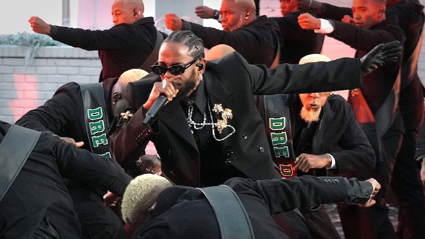 Kendrick Lamar performs at the 2022 Super Bowl, wearing black suit and black glasses with gold jewellry