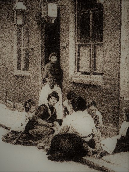 A group of women and children sitting outside on a London street, dressed in Victorian garb