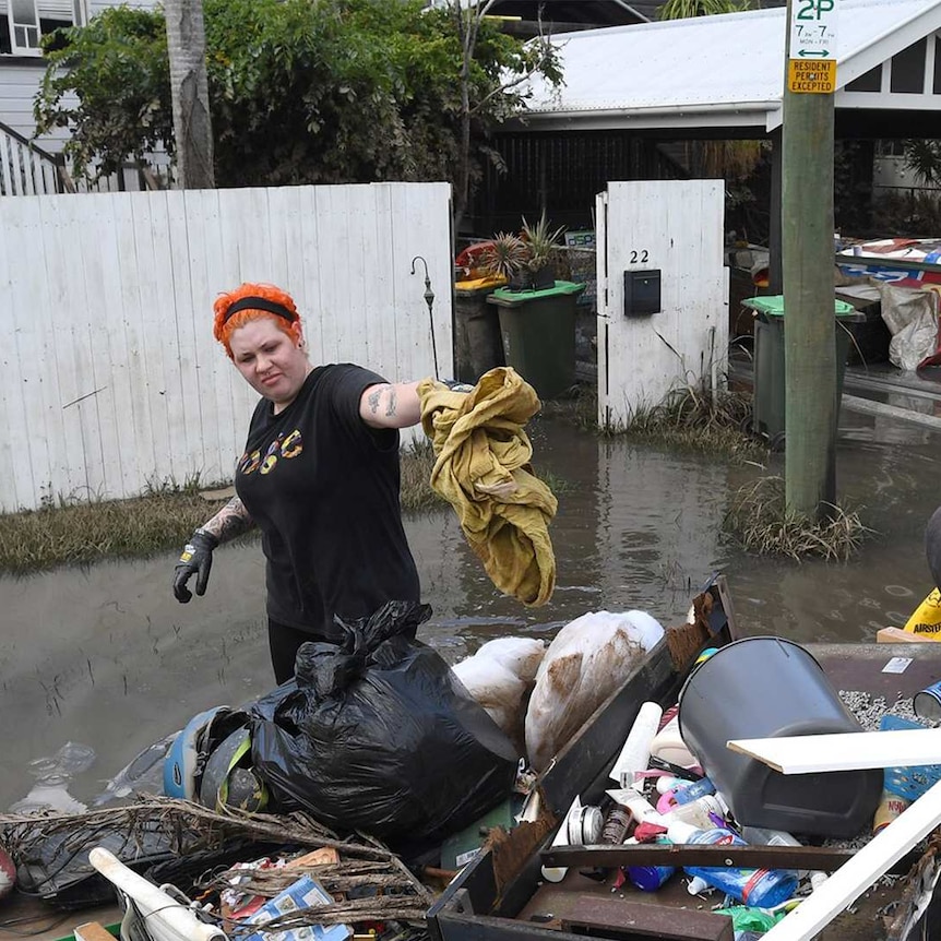 A woman throwing items onto a large pile of rubbish outside her home in low-level flood water.